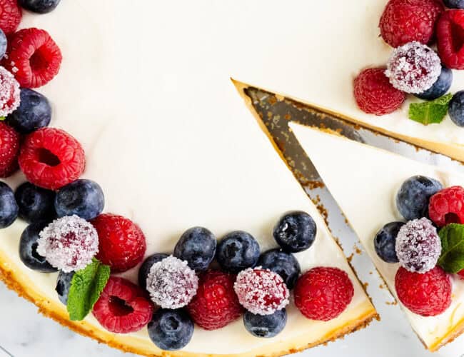 A New York cheesecake topped with fresh raspberries, blueberries, and mint leaves. A slice has been cut from the cake, revealing a creamy interior. The cake is elegantly displayed on a white marble surface.