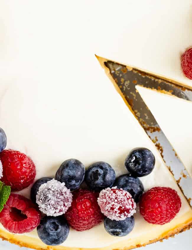 A New York cheesecake topped with fresh raspberries, blueberries, and mint leaves. A slice has been cut from the cake, revealing a creamy interior. The cake is elegantly displayed on a white marble surface.