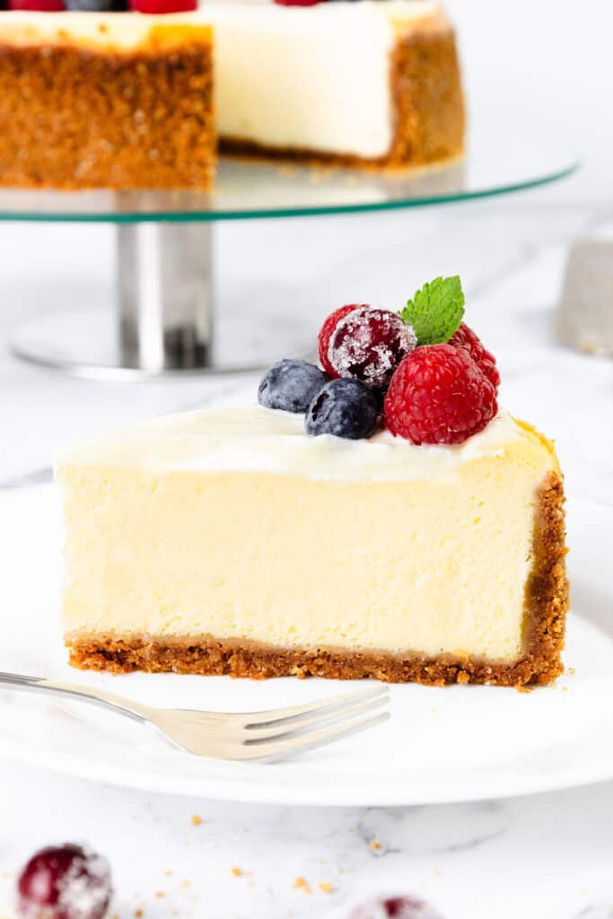 A slice of creamy New York cheesecake with a graham cracker crust is topped with fresh raspberries, blueberries, and a mint leaf on a white plate. A fork rests nearby. Another cheesecake is visible in the background.