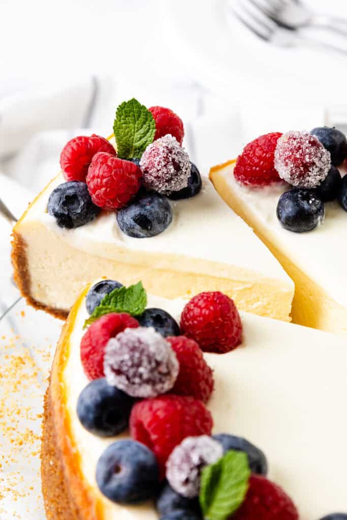 A creamy New York cheesecake topped with sugared berries, including raspberries and blueberries, and fresh mint leaves. A slice of the cheesecake is cut, revealing its smooth texture. The dessert rests on a white surface with forks in the background.