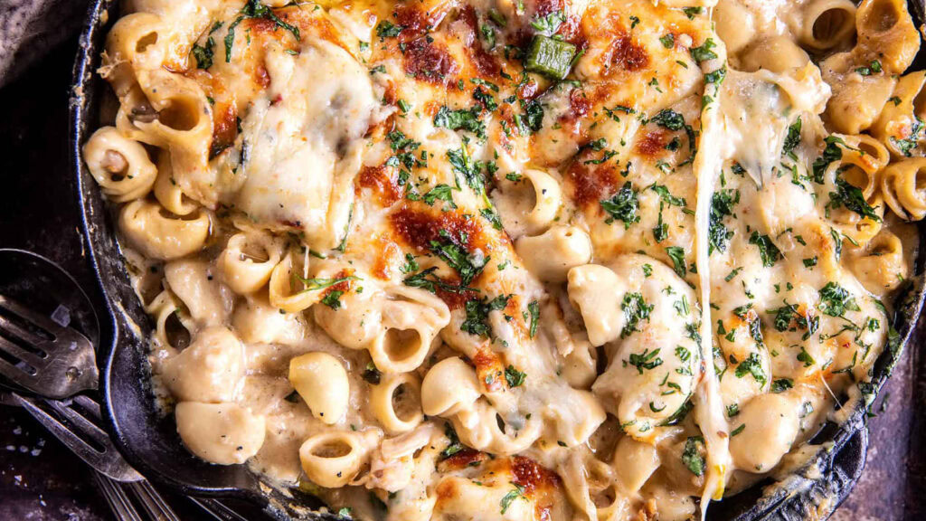 A skillet filled with creamy, cheesy pasta, a perfect example of one pot recipes, is topped with melted cheese and fresh herbs. The shell-shaped pasta boasts a golden-brown crust and hints of green from chopped herbs. Two forks sit beside the skillet, inviting you to indulge.