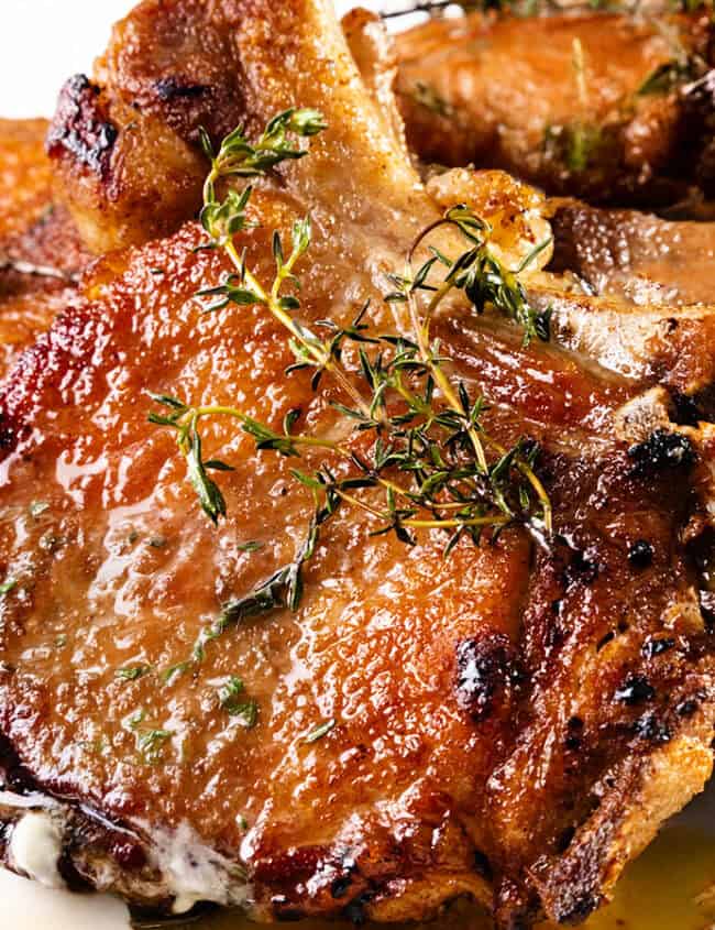 Juicy pan seared pork chops garnished with fresh thyme on a white plate. The chops have a golden-brown crust and are glistening with sauce, highlighting their tenderness and rich flavor.