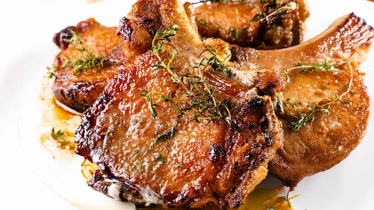 Juicy pan seared pork chops garnished with fresh thyme on a white plate. The chops have a golden-brown crust and are glistening with sauce, highlighting their tenderness and rich flavor.