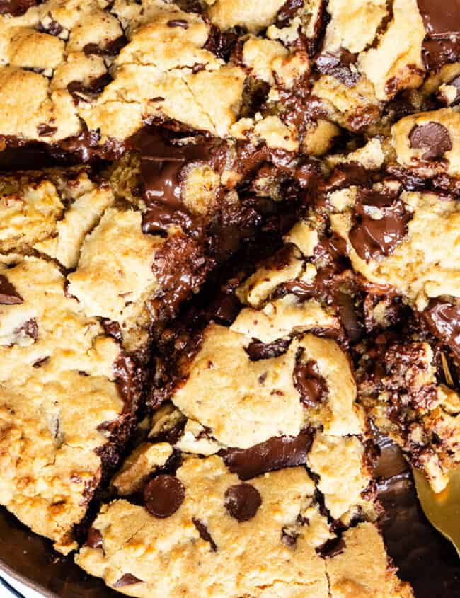 A large, gooey chocolate chip cookie sliced into triangular pieces rests on a cooling rack. A metal spatula is partially under one slice, ready to serve this delectable dessert. Chocolate chunks are visibly melted and spread throughout, inviting you to indulge in its sweetness.