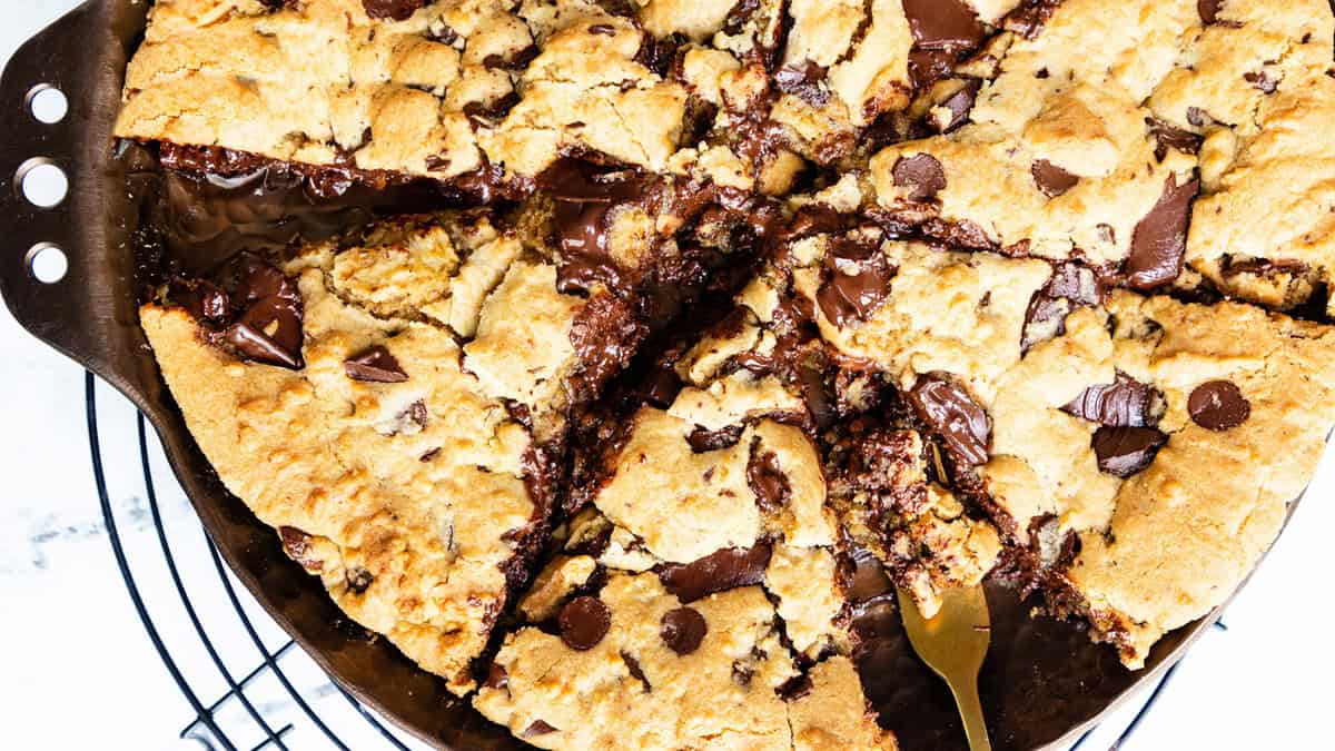 A large, gooey chocolate chip cookie sliced into triangular pieces rests on a cooling rack. A metal spatula is partially under one slice, ready to serve this delectable dessert. Chocolate chunks are visibly melted and spread throughout, inviting you to indulge in its sweetness.