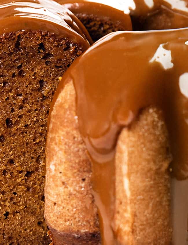 Close-up of a Bundt cake drizzled with glossy caramel sauce, the dessert glistening enticingly. One slice is partially removed, revealing the rich, moist texture of the cake. The indulgent treat rests elegantly on a metal surface.