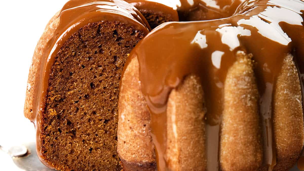 Close-up of a Bundt cake drizzled with glossy caramel sauce, the dessert glistening enticingly. One slice is partially removed, revealing the rich, moist texture of the cake. The indulgent treat rests elegantly on a metal surface.