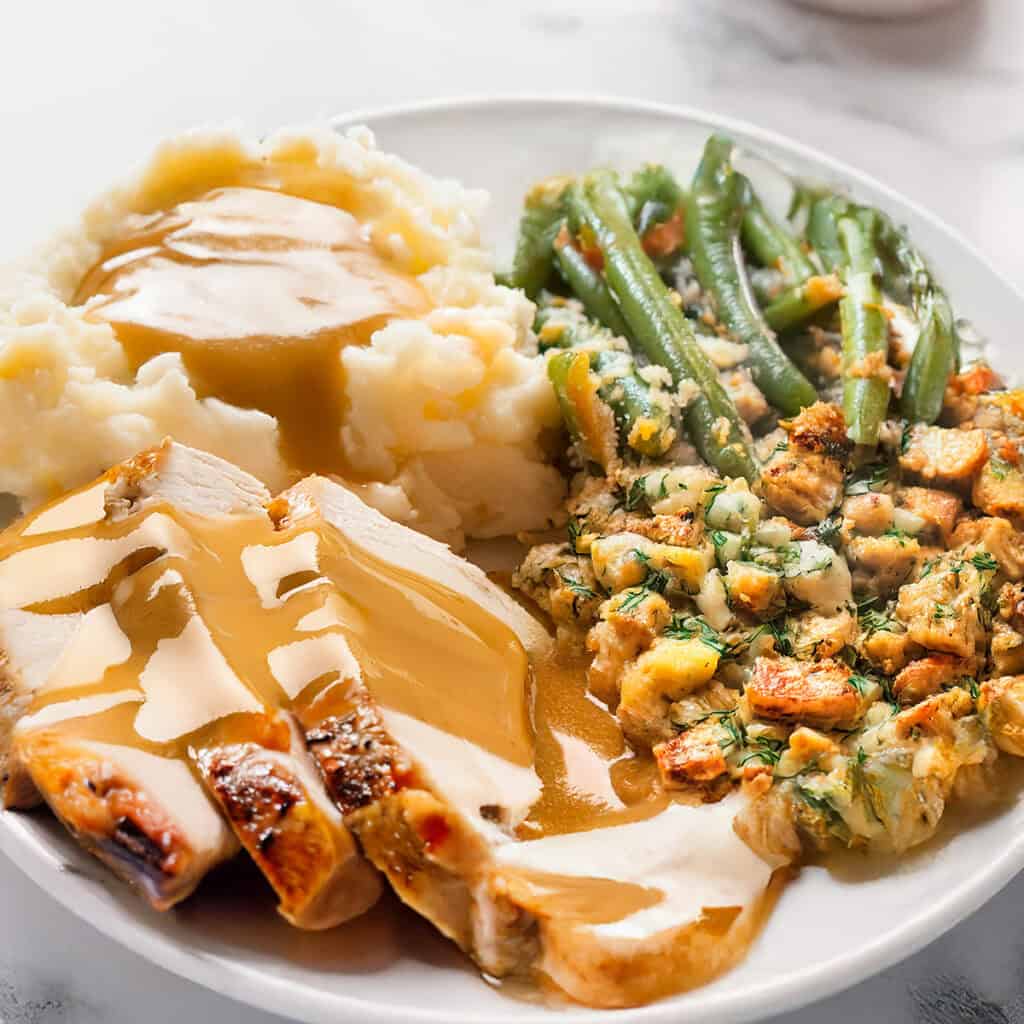 A plate featuring slow roasted turkey slices with gravy, creamy mashed potatoes topped with gravy, green beans, and a serving of seasoned stuffing. The food is arranged neatly on a white plate.