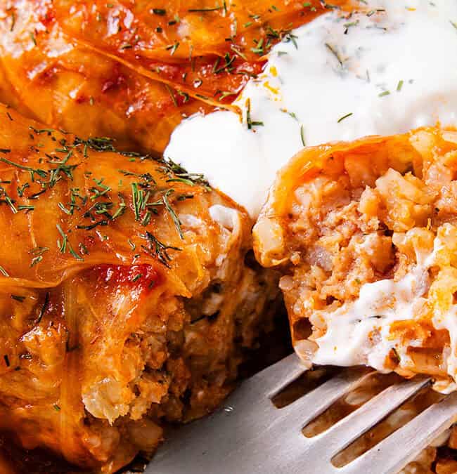 Close-up of stuffed cabbage rolls on a plate, topped with creamy yogurt sauce and garnished with herbs. A fork is cutting into one roll, revealing a filling of rice and ground meat.