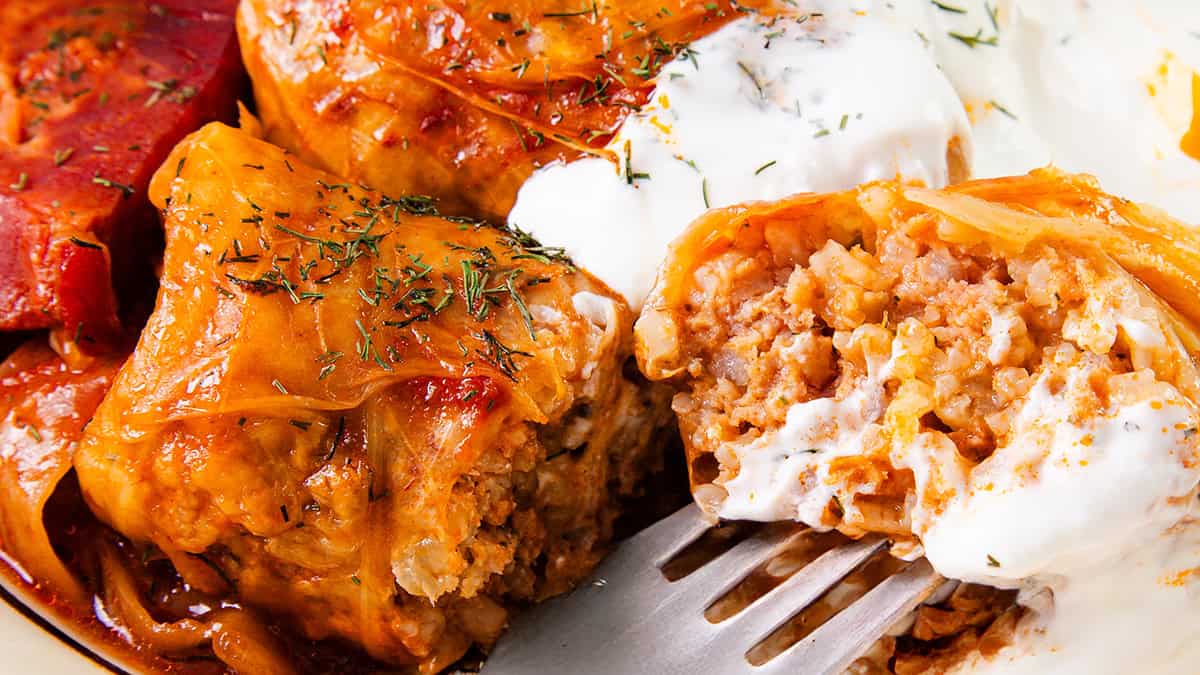 Close-up of stuffed cabbage rolls on a plate, topped with creamy yogurt sauce and garnished with herbs. A fork is cutting into one roll, revealing a filling of rice and ground meat.