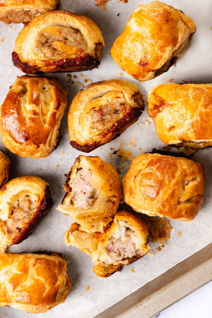 Golden-brown sausage rolls with flaky pastry are scattered on a parchment-lined baking sheet. This delicious recipe features some whole rolls, while a few are broken open, revealing the seasoned sausage filling. The rolls look freshly baked and crisp.