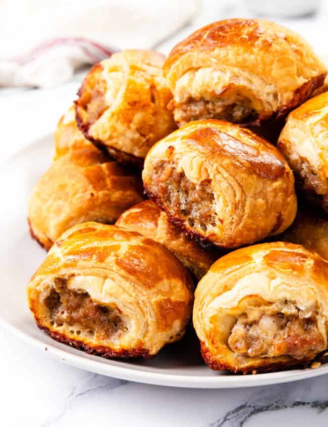 A plate of homemade, golden-brown sausage rolls with flaky pastry is stacked neatly on a white plate. The background features a light-colored cloth and a small bowl, slightly out of focus.