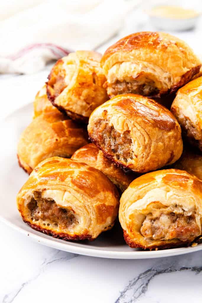 A plate of homemade, golden-brown sausage rolls with flaky pastry is stacked neatly on a white plate. The background features a light-colored cloth and a small bowl, slightly out of focus.