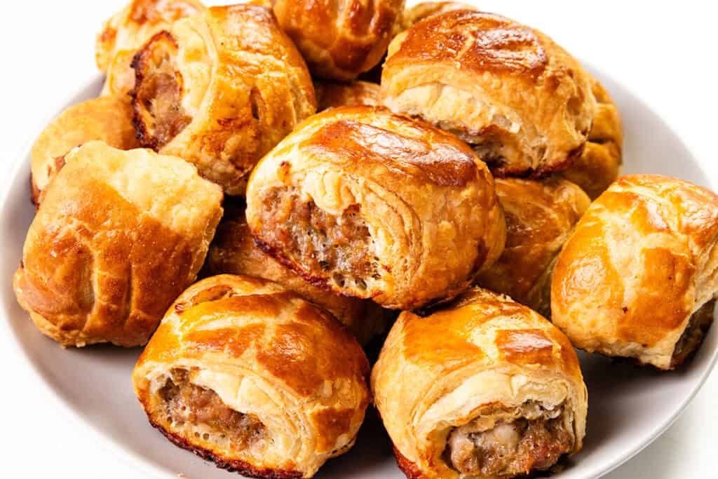 A plate filled with golden-brown homemade sausage rolls. The rolls are flaky and baked to a crisp, revealing a savory filling. They are arranged in a pile on a white plate, with a glossy sheen on the pastry.