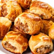 A plate filled with golden-brown homemade sausage rolls. The rolls are flaky and baked to a crisp, revealing a savory filling. They are arranged in a pile on a white plate, with a glossy sheen on the pastry.