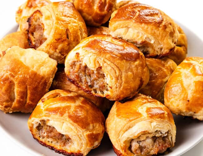 A plate filled with golden-brown homemade sausage rolls. The rolls are flaky and baked to a crisp, revealing a savory filling. They are arranged in a pile on a white plate, with a glossy sheen on the pastry.