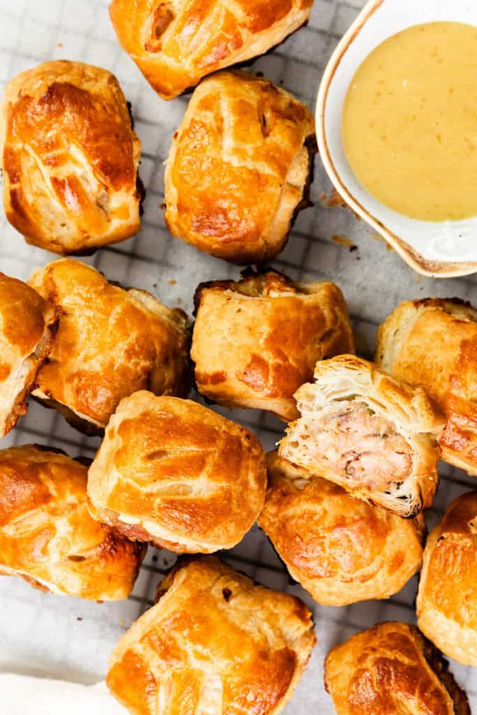 Golden brown homemade sausage rolls arranged on a cooling rack with a small bowl of mustard dip on the side. One roll is cut open, revealing its delicious filling, inviting you to explore the full recipe and savor every bite.