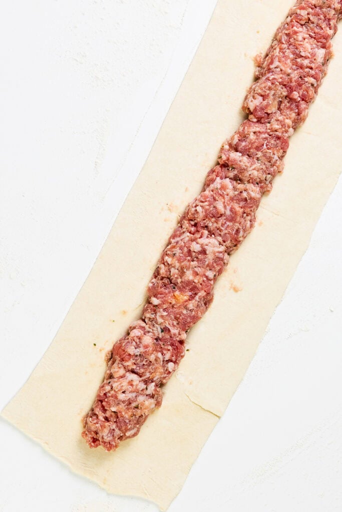 A strip of homemade sausage filling rests on a rectangular sheet of pastry, ready for wrapping into delicious sausage rolls. The mixture contains minced meat and herbs, set against a light, neutral surface.