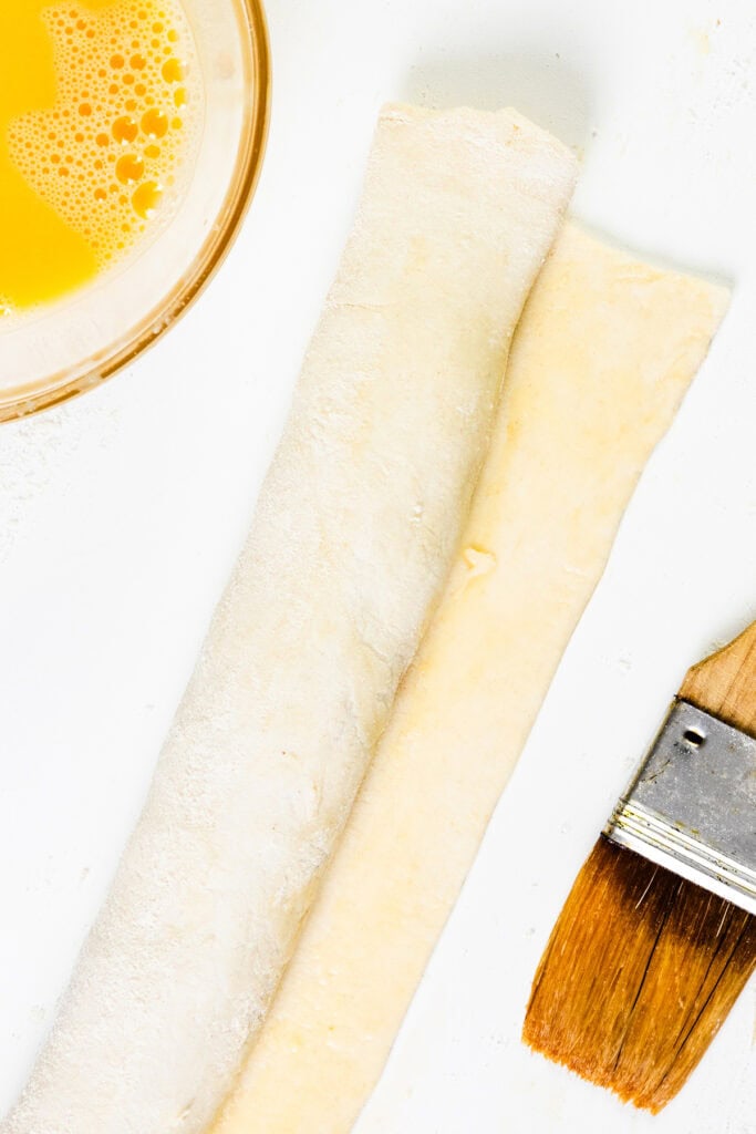 Top view of two rolled-out sheets of dough on a white surface, perfect for crafting homemade sausage rolls. A bowl of beaten egg sits nearby, with a pastry brush positioned to the right, ready for use.