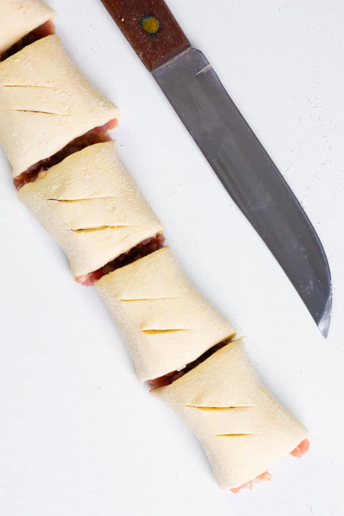 Homemade unbaked sausage rolls with diagonal slits, filled with a savory meat mixture, lie on a white surface next to a large kitchen knife.