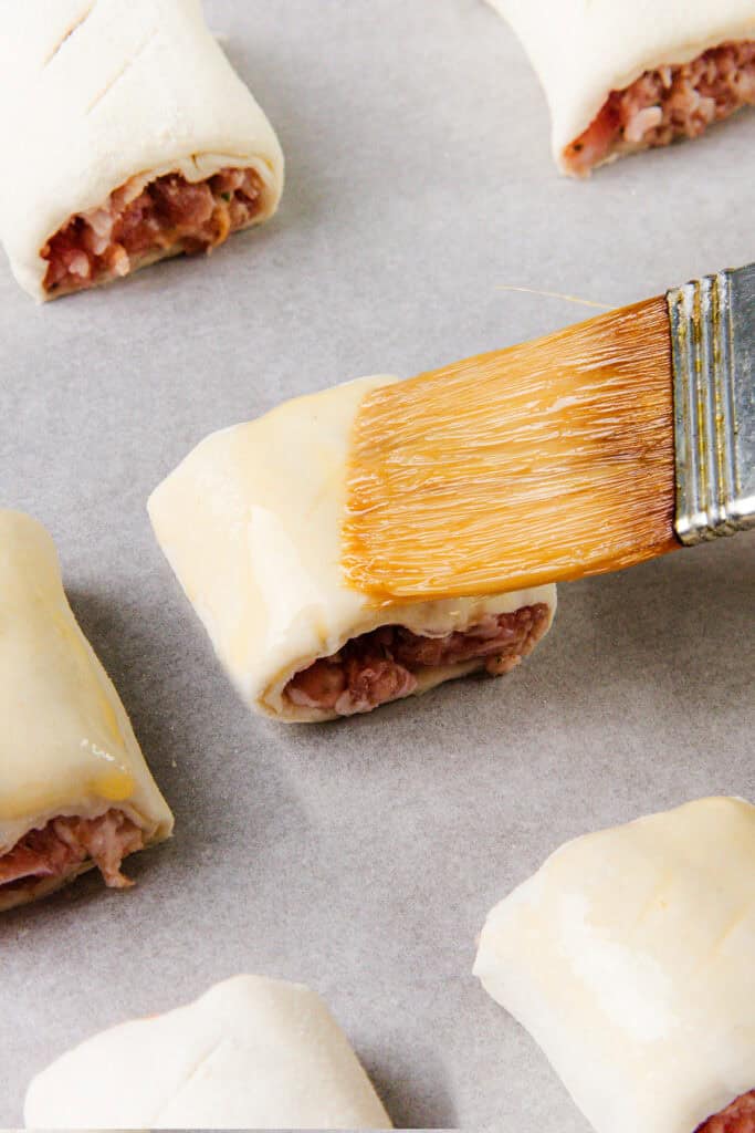 A close-up of a pastry brush applying egg wash to homemade sausage rolls on parchment paper. The uncooked rolls reveal the visible sausage filling inside the lightly scored dough, showcasing a delightful recipe in the making.