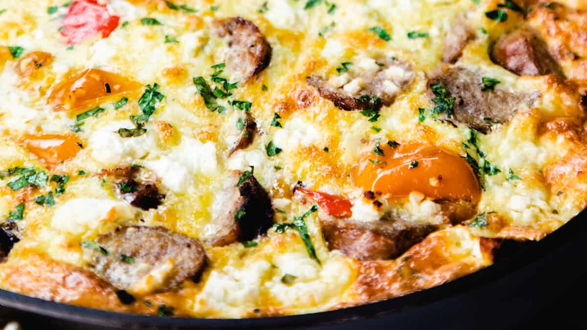 A close-up of a freshly baked frittata with browned sausage slices, red and yellow bell peppers, and a golden-brown cheese topping, garnished with finely chopped parsley.