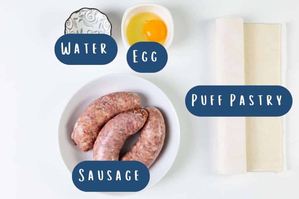 Image of ingredients labeled for homemade sausage rolls: a white bowl with raw sausages, an egg in a small white bowl, a decorative bowl with water, and a roll of puff pastry on a pristine white surface.