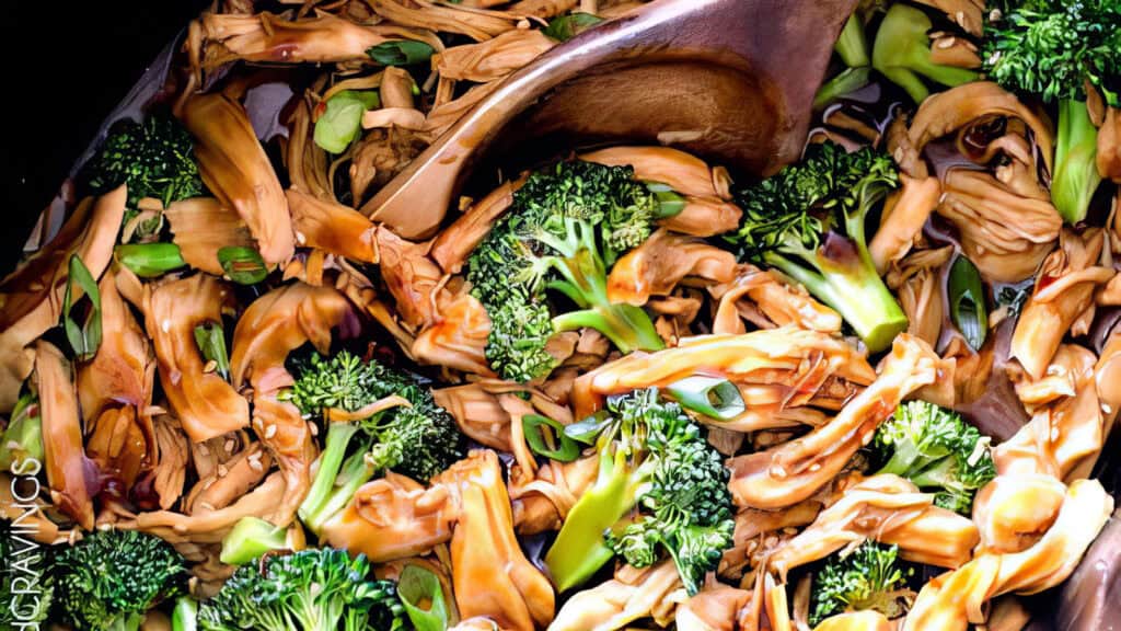 A vibrant stir-fry with broccoli and shredded jackfruit, mixed with green onions in a pan. A wooden spoon stirs the colorful blend, surrounded by an array of greens and the rich golden hue of the jackfruit—a quick delight amid slow cooker recipes.