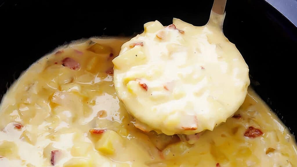 A ladle scooping creamy potato soup with visible chunks of potatoes and bacon from a slow cooker. The thick, rich soup boasts a smooth yellowish color, perfect for warming up on a chilly day.