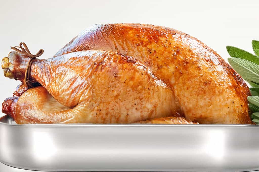 A golden-brown slow roasted turkey sits in a metal roasting pan, garnished with fresh sage leaves. The turkey's crispy skin glistens, suggesting a perfectly cooked, juicy roast.