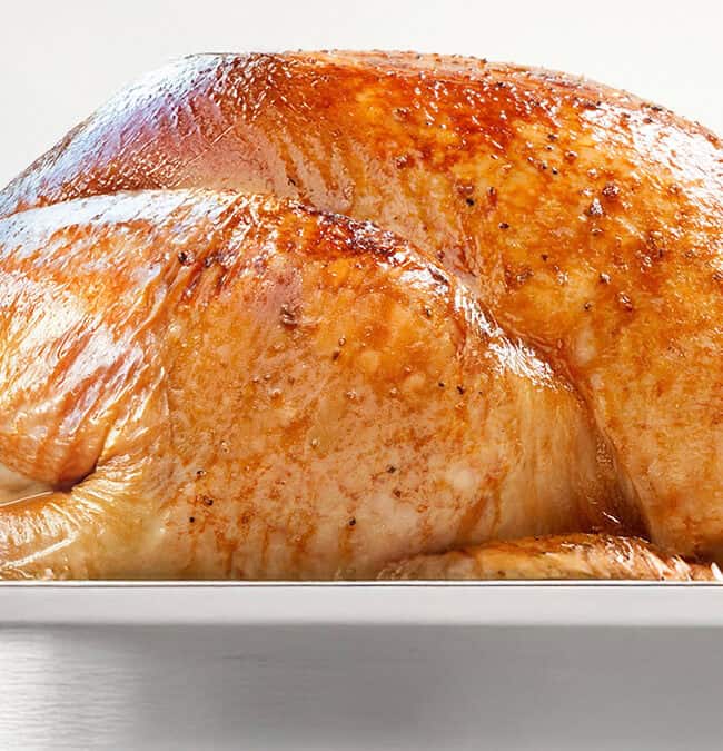 Golden-brown roasted turkey in a silver roasting pan, garnished with fresh sage leaves on the side. The turkey has crispy, seasoned skin, and is tied at the legs with twine.