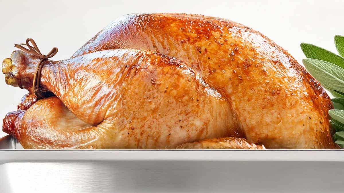 Golden-brown roasted turkey in a silver roasting pan, garnished with fresh sage leaves on the side. The turkey has crispy, seasoned skin, and is tied at the legs with twine.