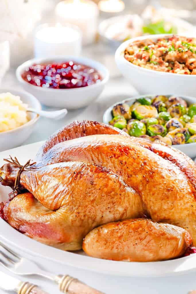 A slow-roasted turkey on a white platter is surrounded by side dishes, including mashed potatoes, Brussels sprouts, and cranberry sauce. Lit candles in the background create a warm, festive ambiance that perfectly complements any turkey recipe.