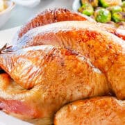 A golden-brown, slow-roasted turkey graces a white platter, garnished with fresh herbs. In the background, a bowl of Brussels sprouts and a dish of mashed potatoes can be seen. The turkey's skin is crispy and shiny, embodying the essence of an overnight roasted masterpiece.