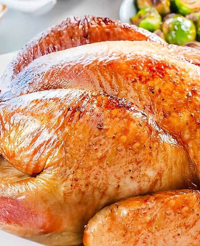 A golden-brown, slow-roasted turkey graces a white platter, garnished with fresh herbs. In the background, a bowl of Brussels sprouts and a dish of mashed potatoes can be seen. The turkey's skin is crispy and shiny, embodying the essence of an overnight roasted masterpiece.