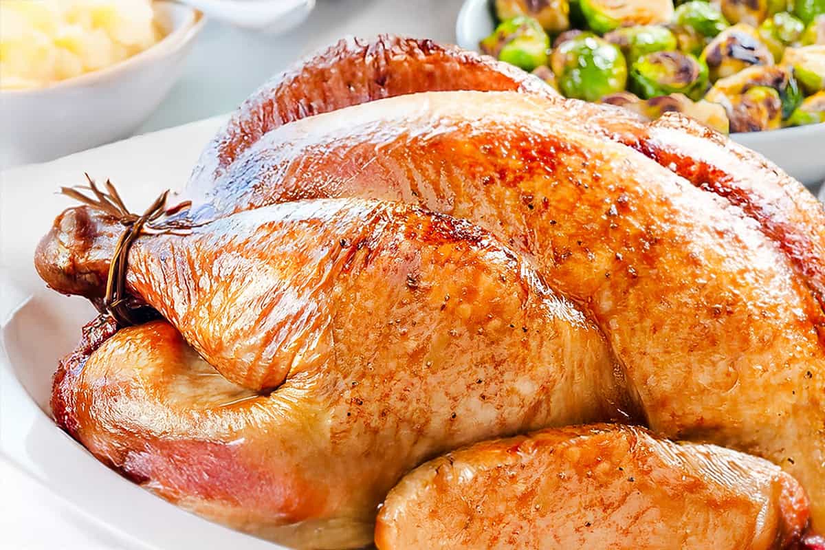 A golden-brown, slow-roasted turkey graces a white platter, garnished with fresh herbs. In the background, a bowl of Brussels sprouts and a dish of mashed potatoes can be seen. The turkey's skin is crispy and shiny, embodying the essence of an overnight roasted masterpiece.