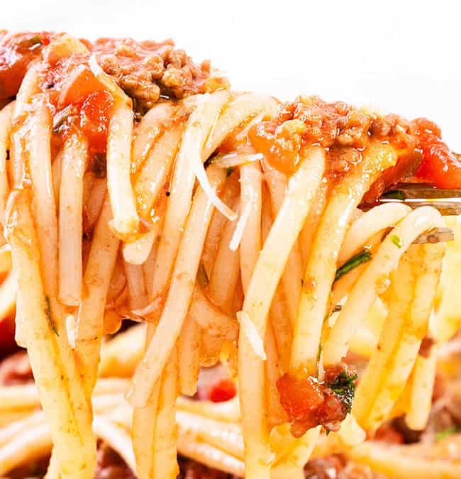 A fork holds a portion of spaghetti topped with tomato sauce, ground meat, and herbs. The background shows more of the pasta dish.