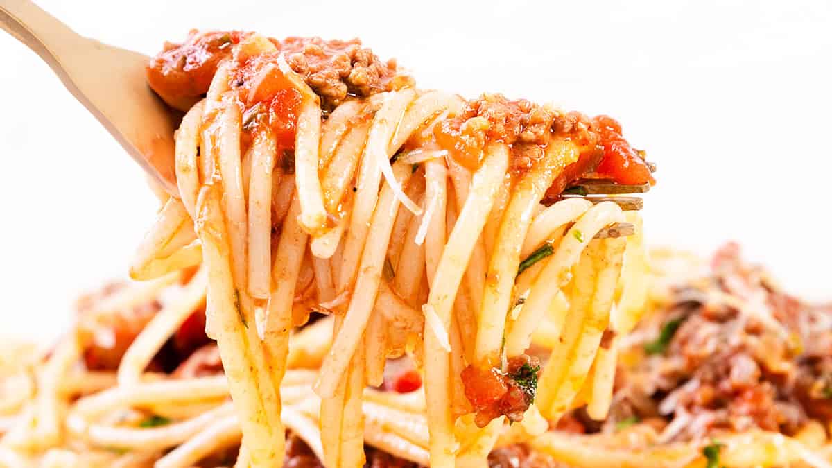 A fork holds a portion of spaghetti topped with tomato sauce, ground meat, and herbs. The background shows more of the pasta dish.