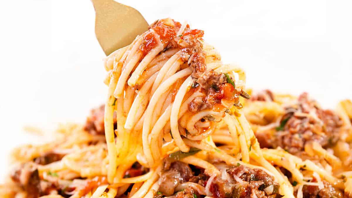 A fork twirls a serving of spaghetti with a rich meat sauce, featuring ground beef, tomatoes, and herbs. The dish is garnished with melted cheese, creating a comforting and savory meal.