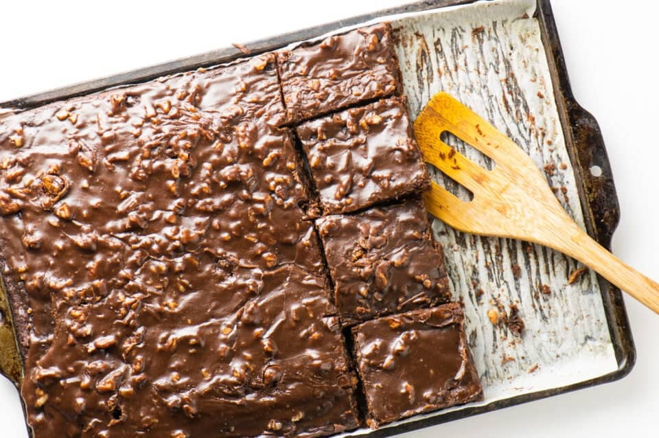 A tray of freshly baked, glazed brownies with a chocolate coating, reminiscent of beloved sheet cake recipes, partially cut into squares. A wooden spatula rests on the baking sheet, hinting at empty spaces where delicious squares were eagerly removed.