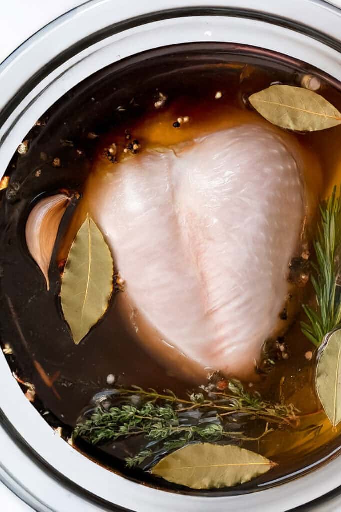 A raw turkey breast submerged in a brining liquid with garlic, bay leaves, black peppercorns, rosemary, and thyme in a white bowl evokes the flavors of a slow-roasted turkey feast.