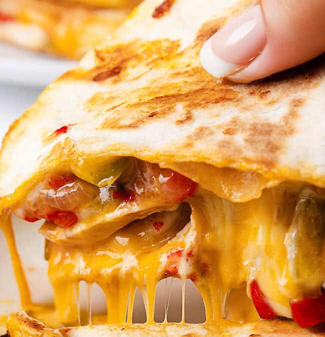 Close-up of a hand holding a cheesy quesadilla with melted cheese, grilled vegetables, and a golden, crispy tortilla, oozing onto a white plate.