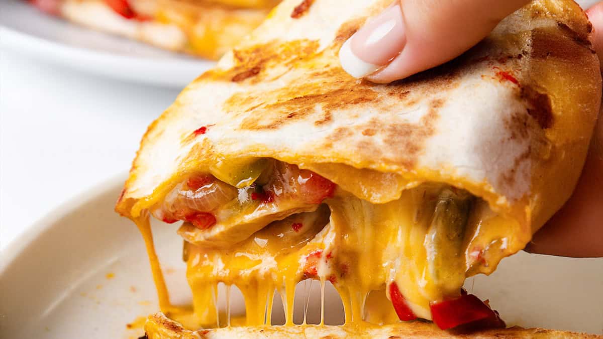 Close-up of a hand holding a cheesy quesadilla with melted cheese, grilled vegetables, and a golden, crispy tortilla, oozing onto a white plate.