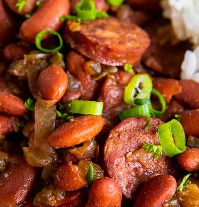 A plate of red beans and sausage stew garnished with sliced green onions, served with white rice. The dish appears savory, with beans and sausage pieces mixed in a flavorful sauce.