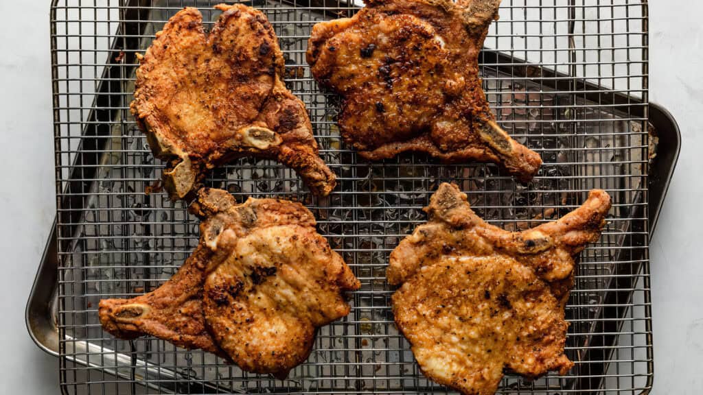 Four breaded and fried pork chops cooling on a wire rack set over a baking sheet, embodying the essence of Southern recipes. The chops are golden brown with a crispy coating reminiscent of classic Southern flavor.