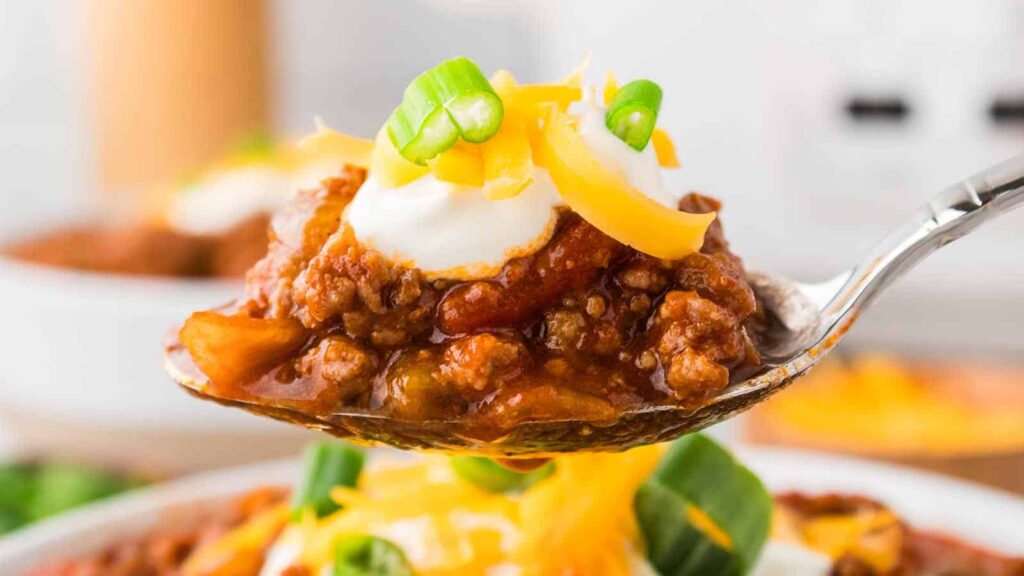 A spoonful of chili topped with sour cream, shredded cheddar cheese, and sliced green onions is held above a bowl. The chili features ground meat and tomato chunks, with a garnish of spinach and cheese visible on the bowl beneath.