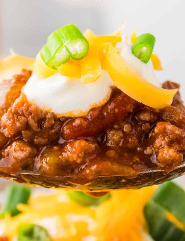 A spoonful of chili topped with sour cream, shredded cheddar cheese, and sliced green onions is held above a bowl. The chili features ground meat and tomato chunks, with a garnish of spinach and cheese visible on the bowl beneath.