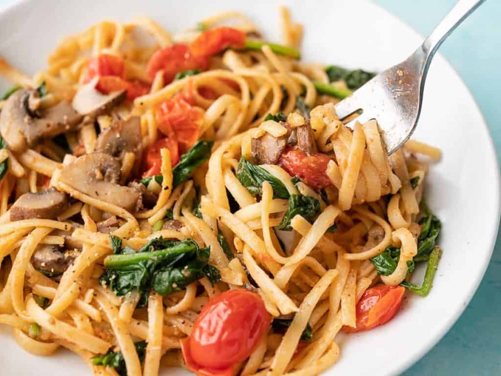 A cozy plate of linguine pasta with mushrooms, spinach, and cherry tomatoes awaits. A fork lifts a portion, inviting you to savor this quick delight. Garnished with herbs and spices, the dish sits against a light blue background—perfect for fall dinners.