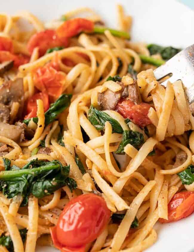 A cozy plate of linguine pasta with mushrooms, spinach, and cherry tomatoes awaits. A fork lifts a portion, inviting you to savor this quick delight. Garnished with herbs and spices, the dish sits against a light blue background—perfect for fall dinners.