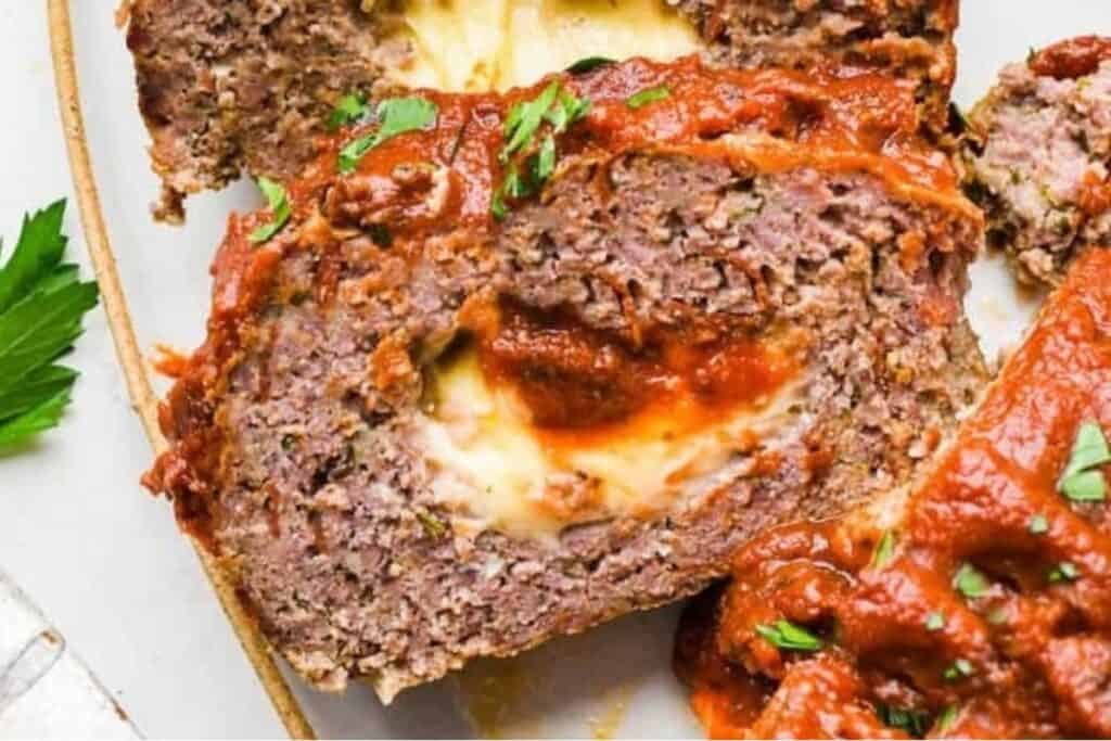 Close-up of a slice of meatloaf stuffed with melted cheese and topped with tomato sauce and herbs. Parsley garnish is visible on the side.